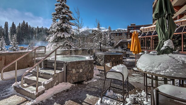 Zona exterior nevada con jacuzzis, una escalera y vistas a árboles y montañas cubiertas de nieve.