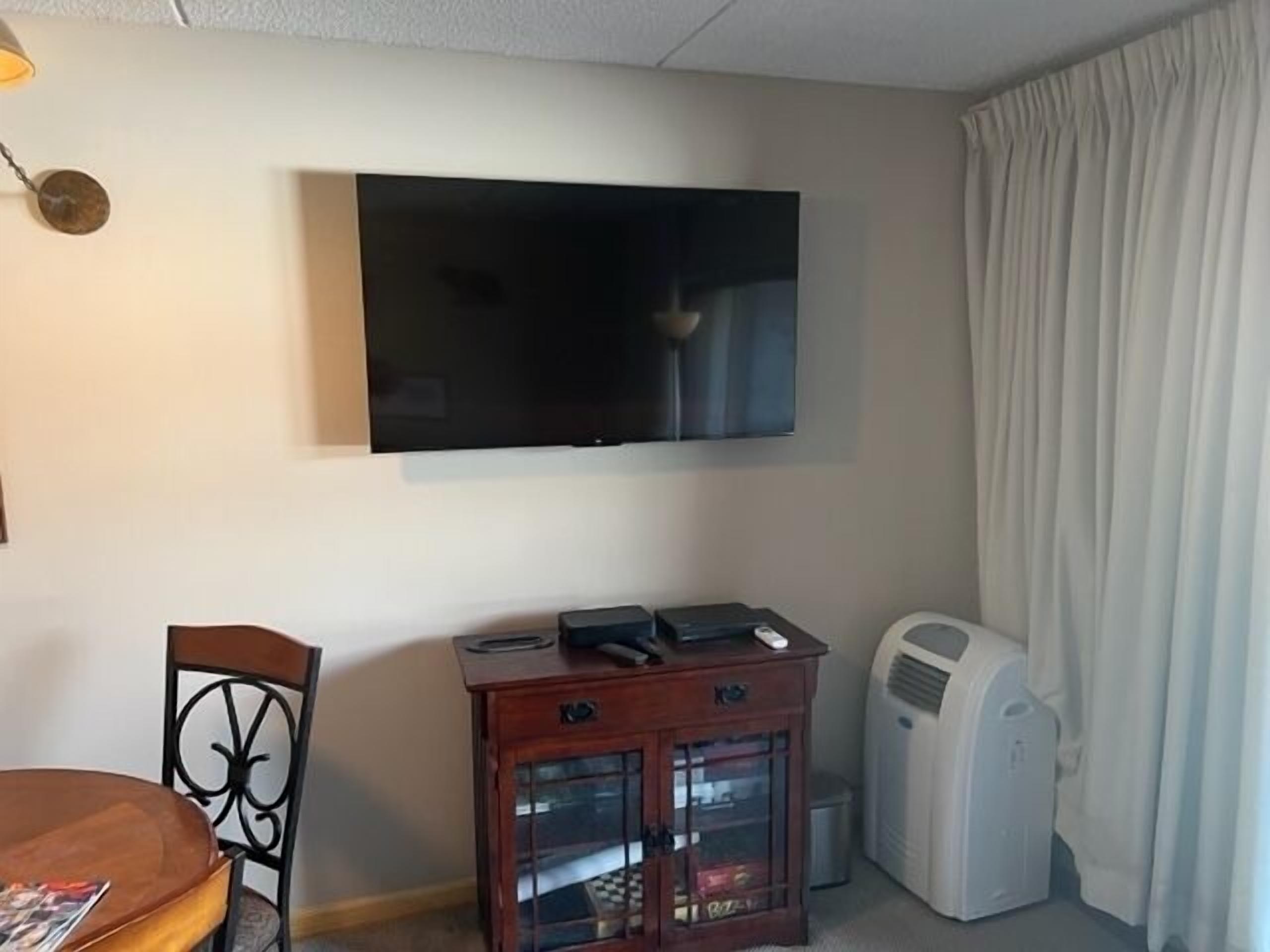A room with a flat-screen TV mounted on the wall, a wooden cabinet with glass doors, a chair, and a table.