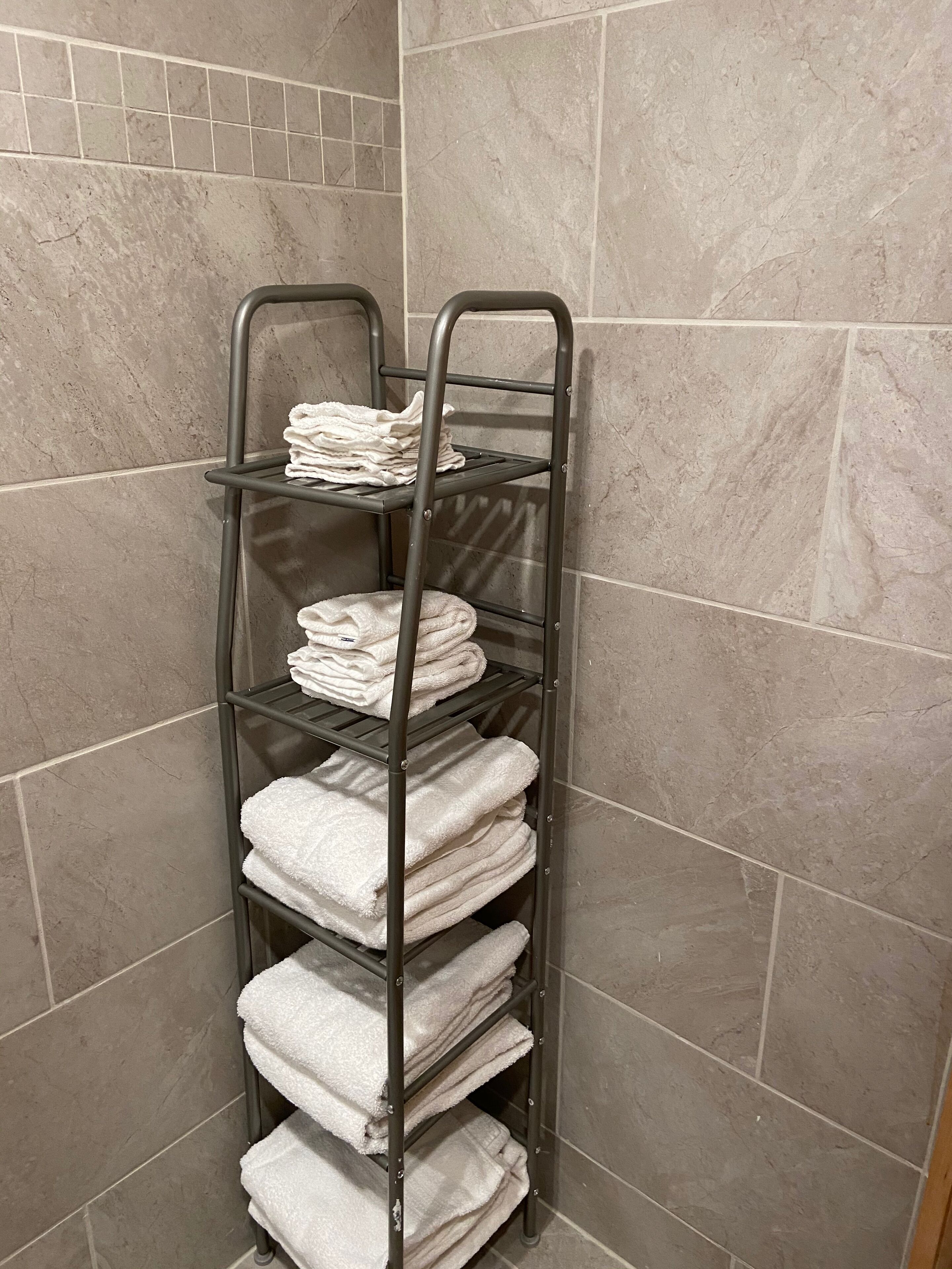 A metal towel rack with multiple shelves holding neatly folded white towels against a tiled wall.