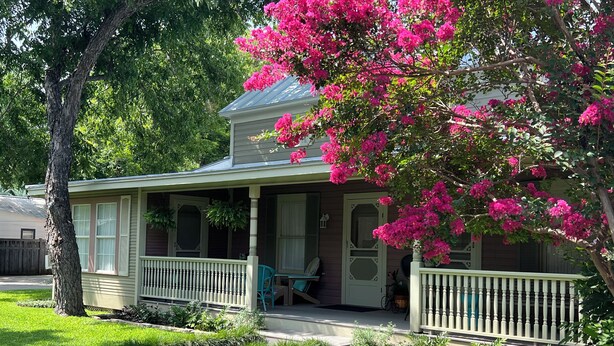 Una casa con porche, un pequeño jardín y un árbol de flores rosadas.