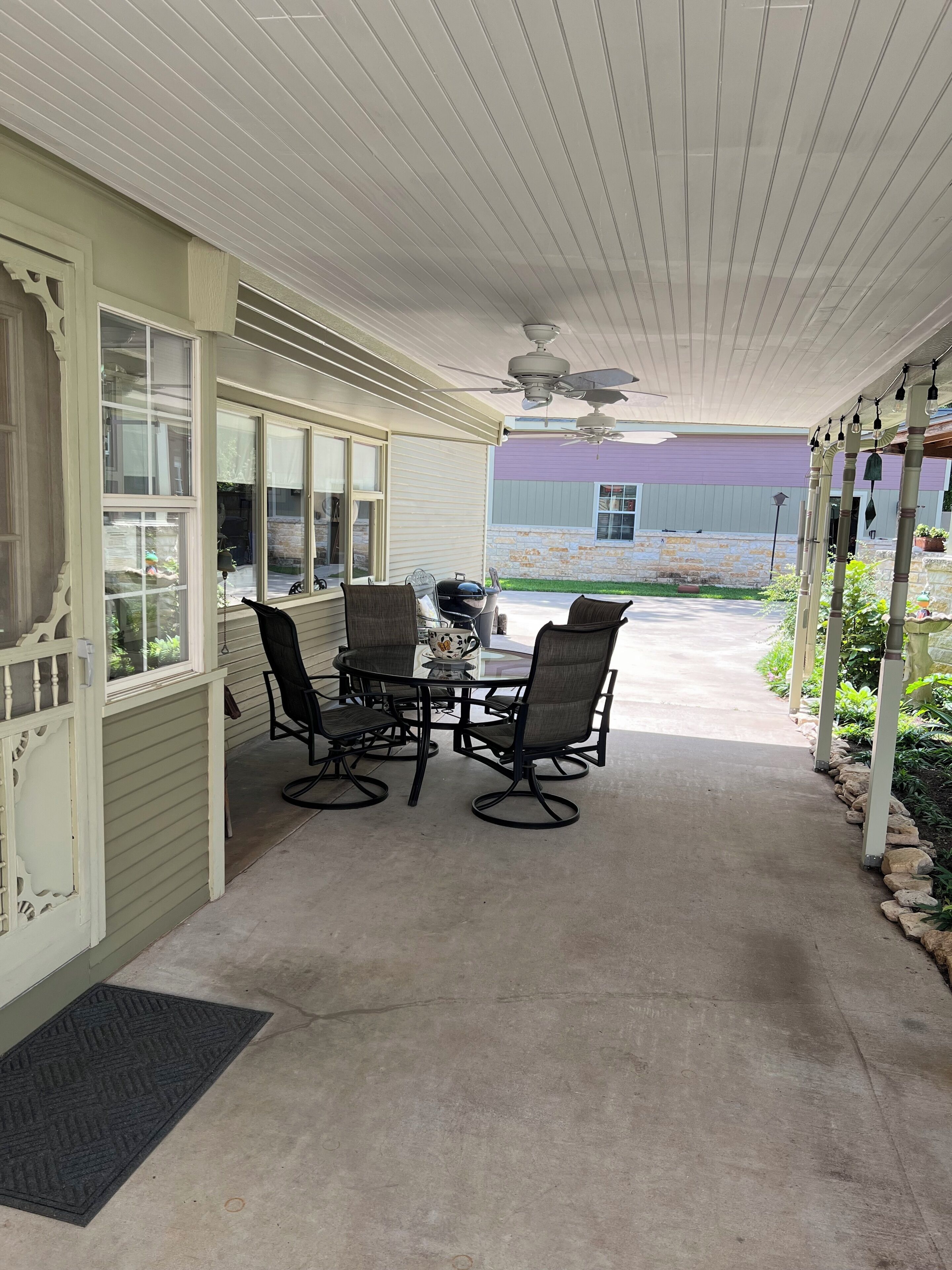 Une terrasse couverte avec une table et des chaises, un ventilateur de plafond et une vue sur un bâtiment en arrière-plan.