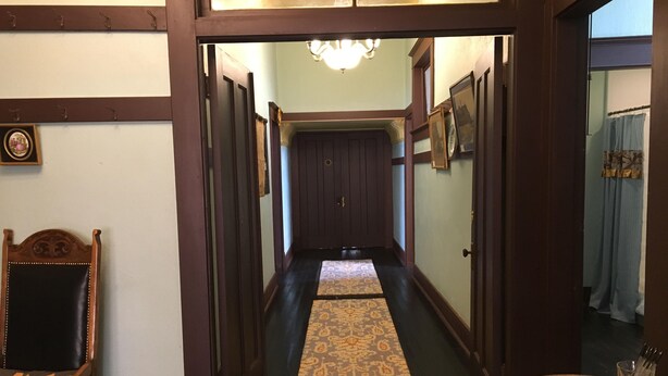 A hallway with a wooden floor, a patterned rug, a chair, a table with a book and glasses, and framed pictures on the walls.