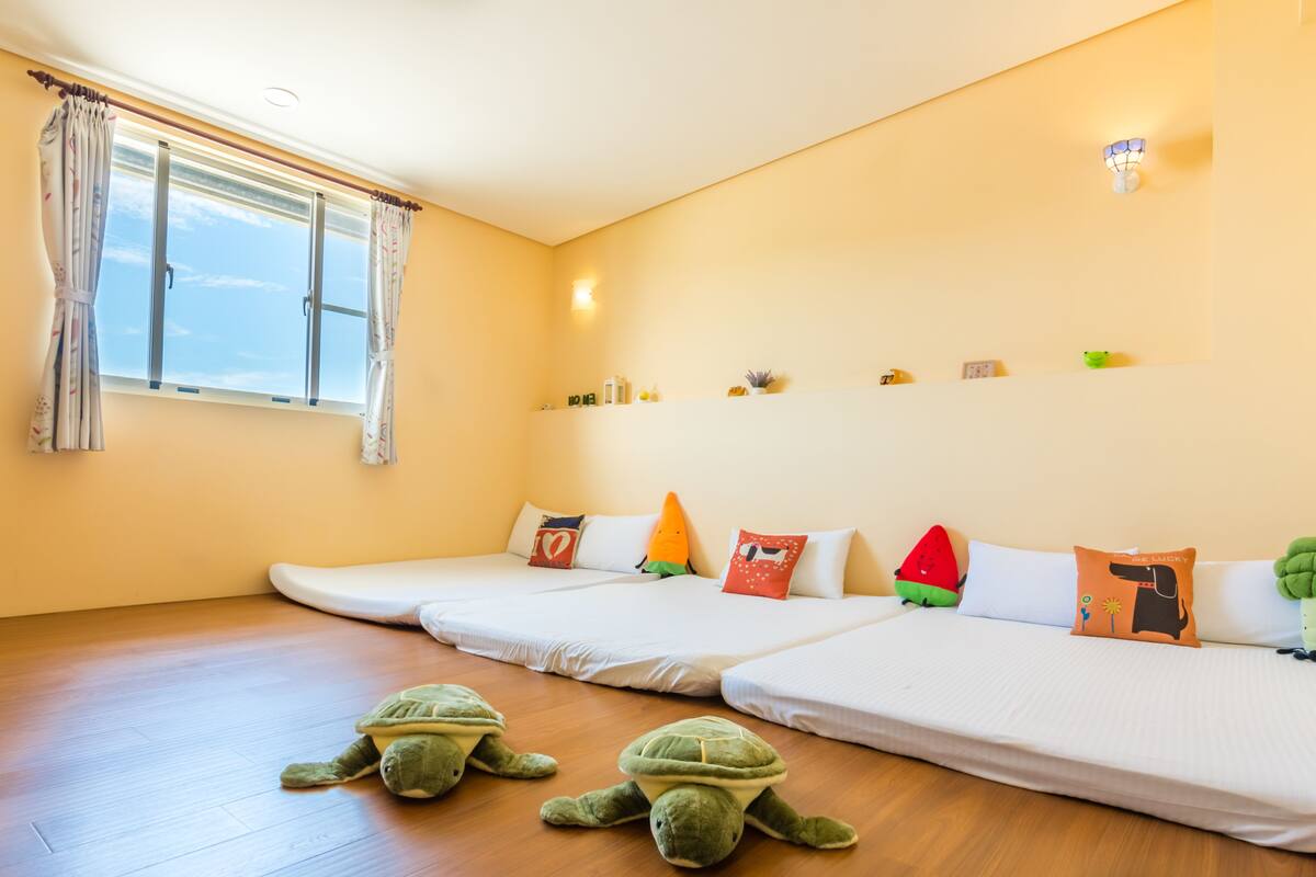 Una habitación con dos camas, una ventana con cortinas, un estante con objetos pequeños y una lámpara fijada a la pared.