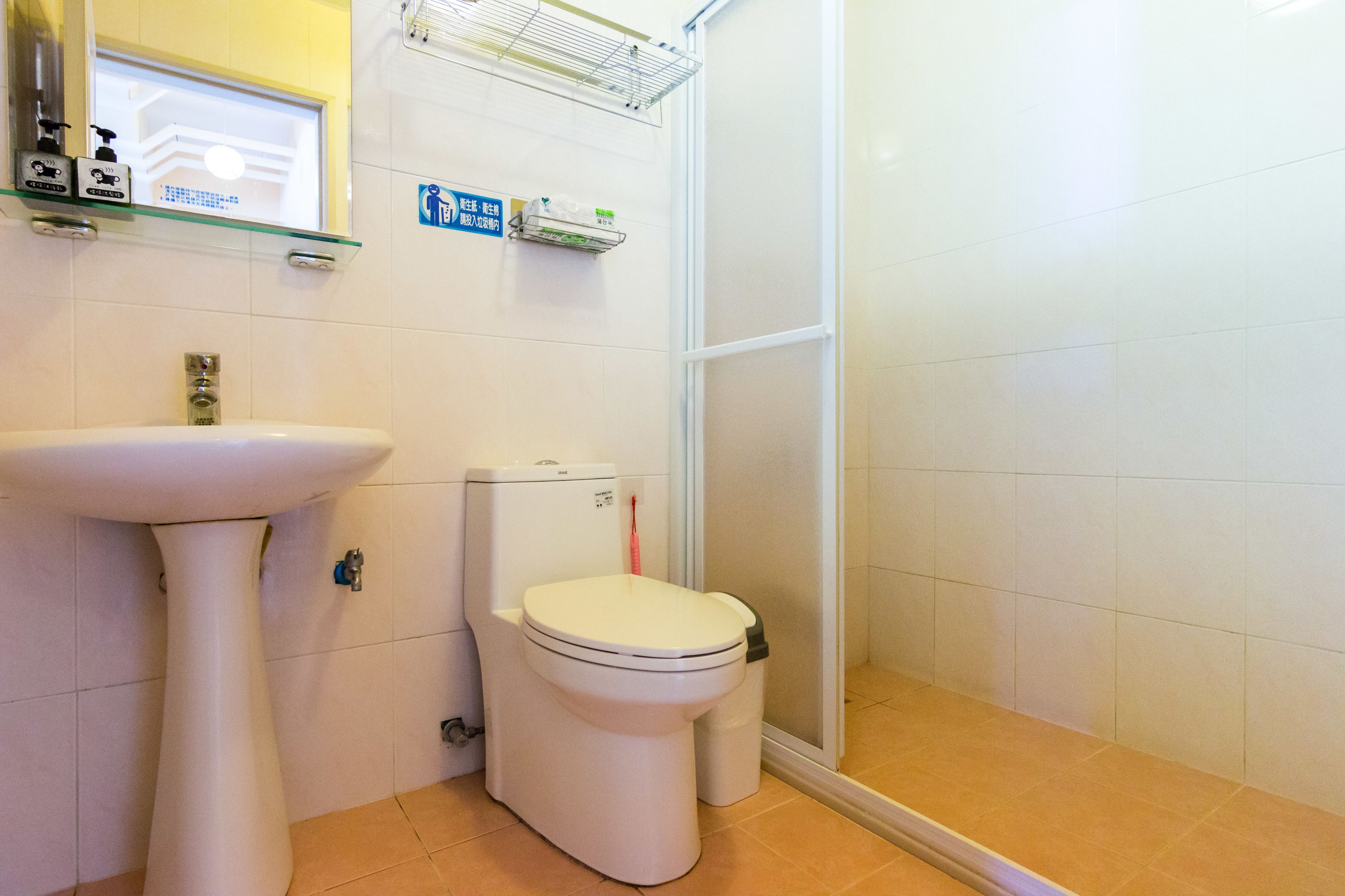 A bathroom with a white sink, toilet, and shower. There are shelves with toiletries and a sign indicating the bathroom's location.