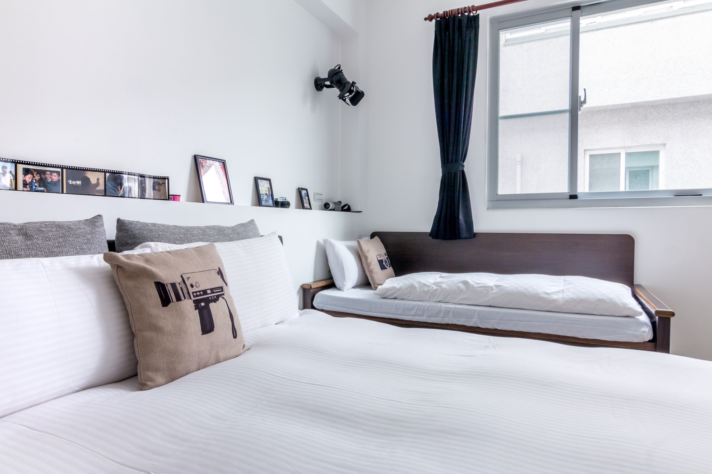 A neatly arranged bedroom with a bed, pillows, a shelf with photos, and a camera on the wall.