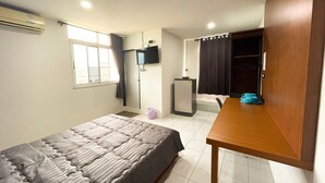 A bedroom with a bed, a wooden table, a TV, a wardrobe, a shower area, and a window. - Sadudee Mansion (Surat Thani)