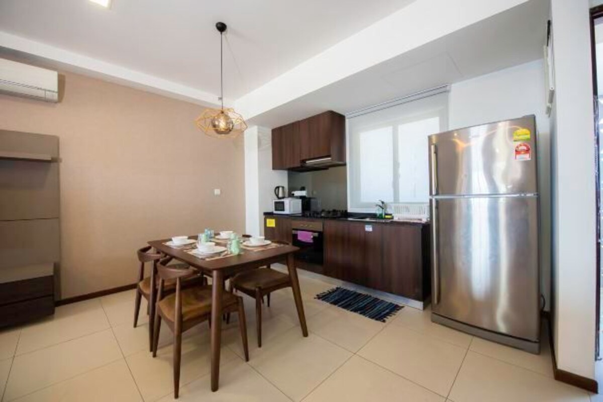A compact kitchen with a stainless steel refrigerator, a microwave, and a sink. There is a dining table with chairs and a hanging light fixture.