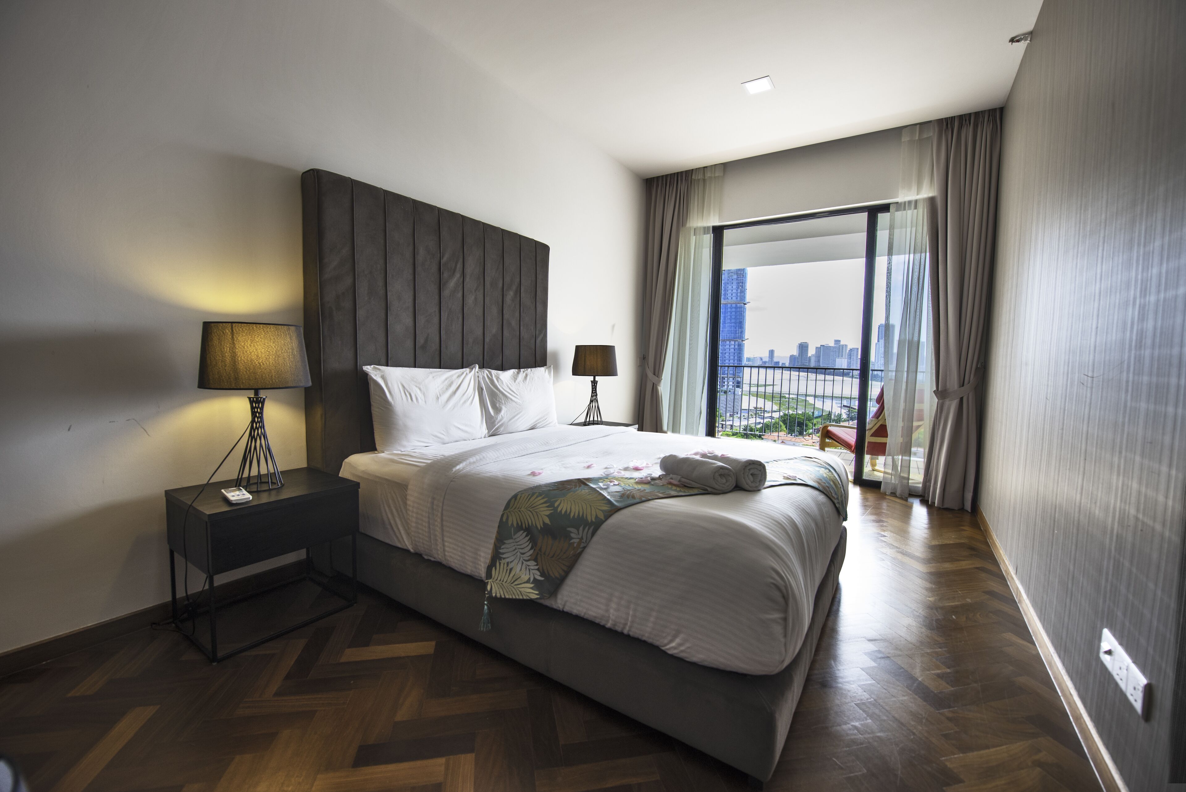 A modern hotel room with a large bed, two bedside lamps, a nightstand, and a view of a cityscape through a large window.