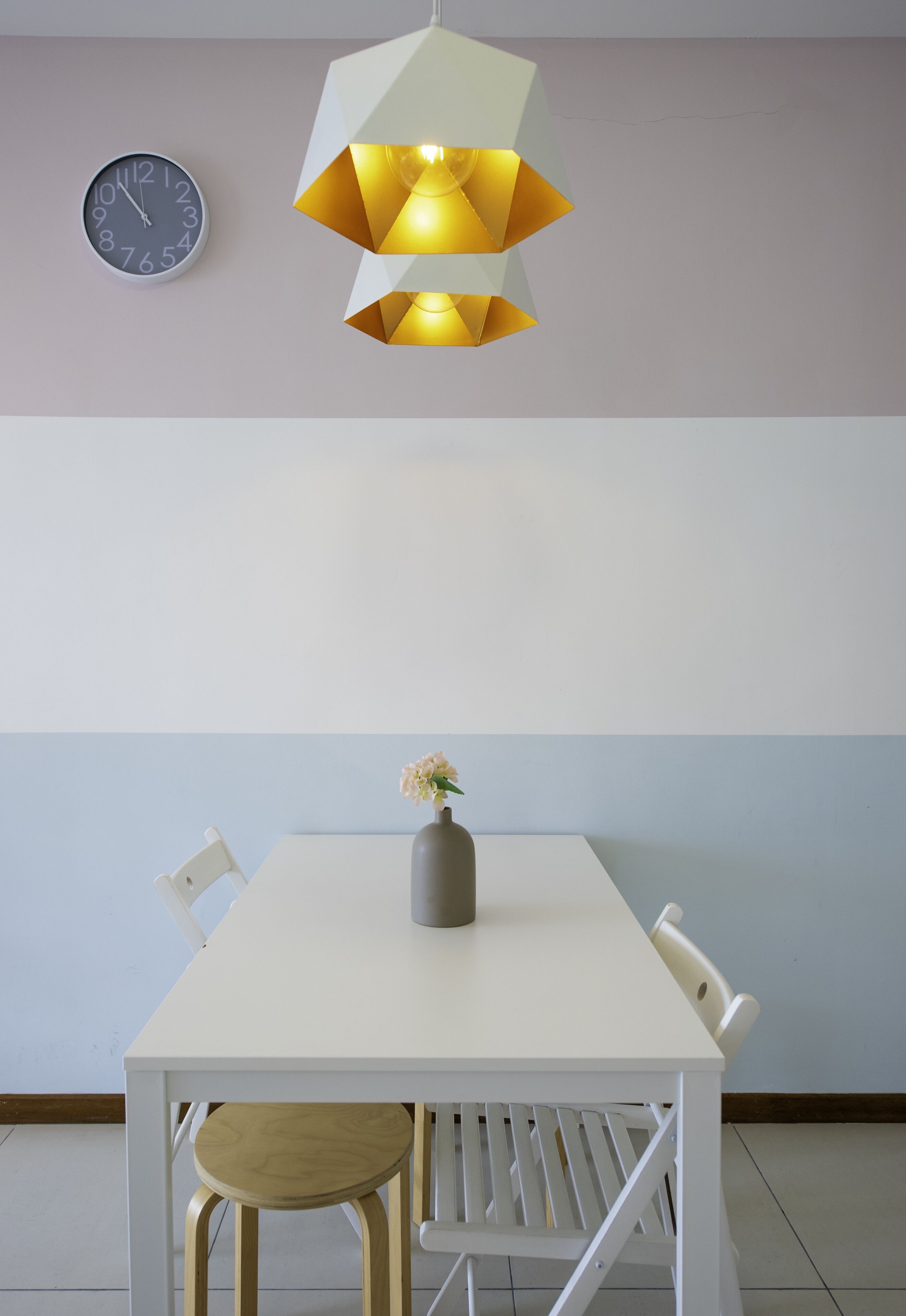 A dining area with a white table, two chairs, a vase with flowers, a wall clock, and a geometric pendant light.