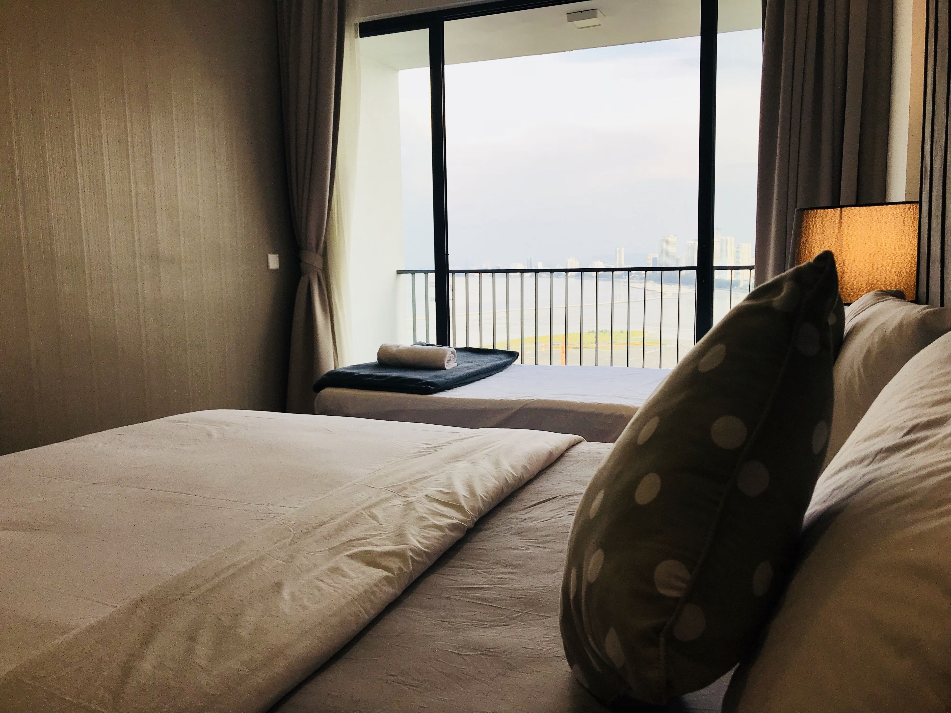 A hotel room with a bed, a small table with a towel, a pillow with polka dots, and a view of a cityscape through a window.