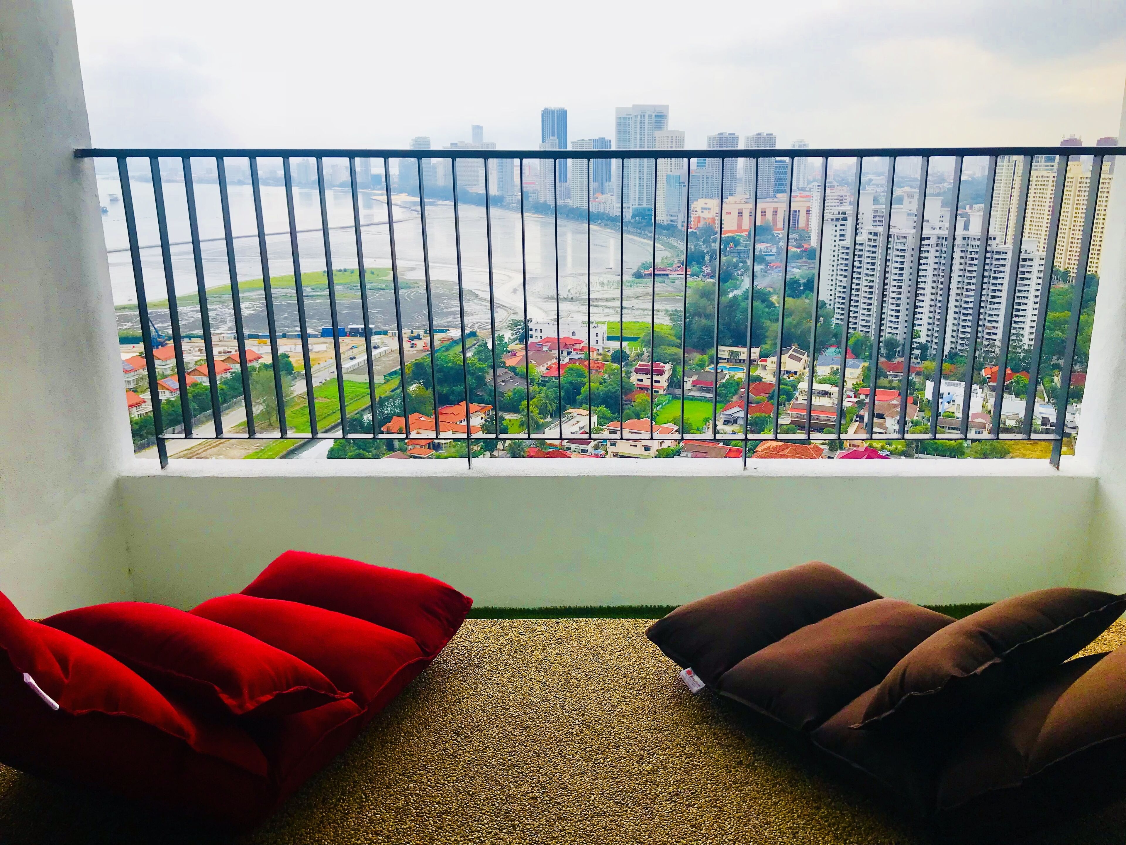 A balcony with a view of a cityscape, including buildings and a river. There are two red and two brown cushions on the floor.