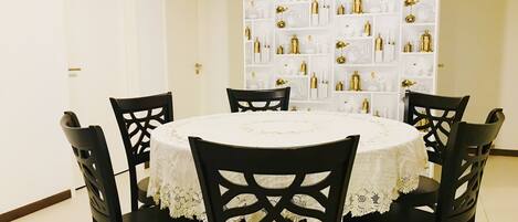 A dining area with a round table covered by a white tablecloth, surrounded by six black chairs with intricate back designs. Behind the table, there is a white shelving unit filled with various kitchenware and decorative items.