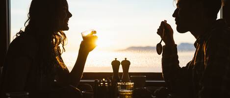 Zwei Personen sitzen an einem Tisch mit Blick auf den Sonnenuntergang durch ein Fenster.