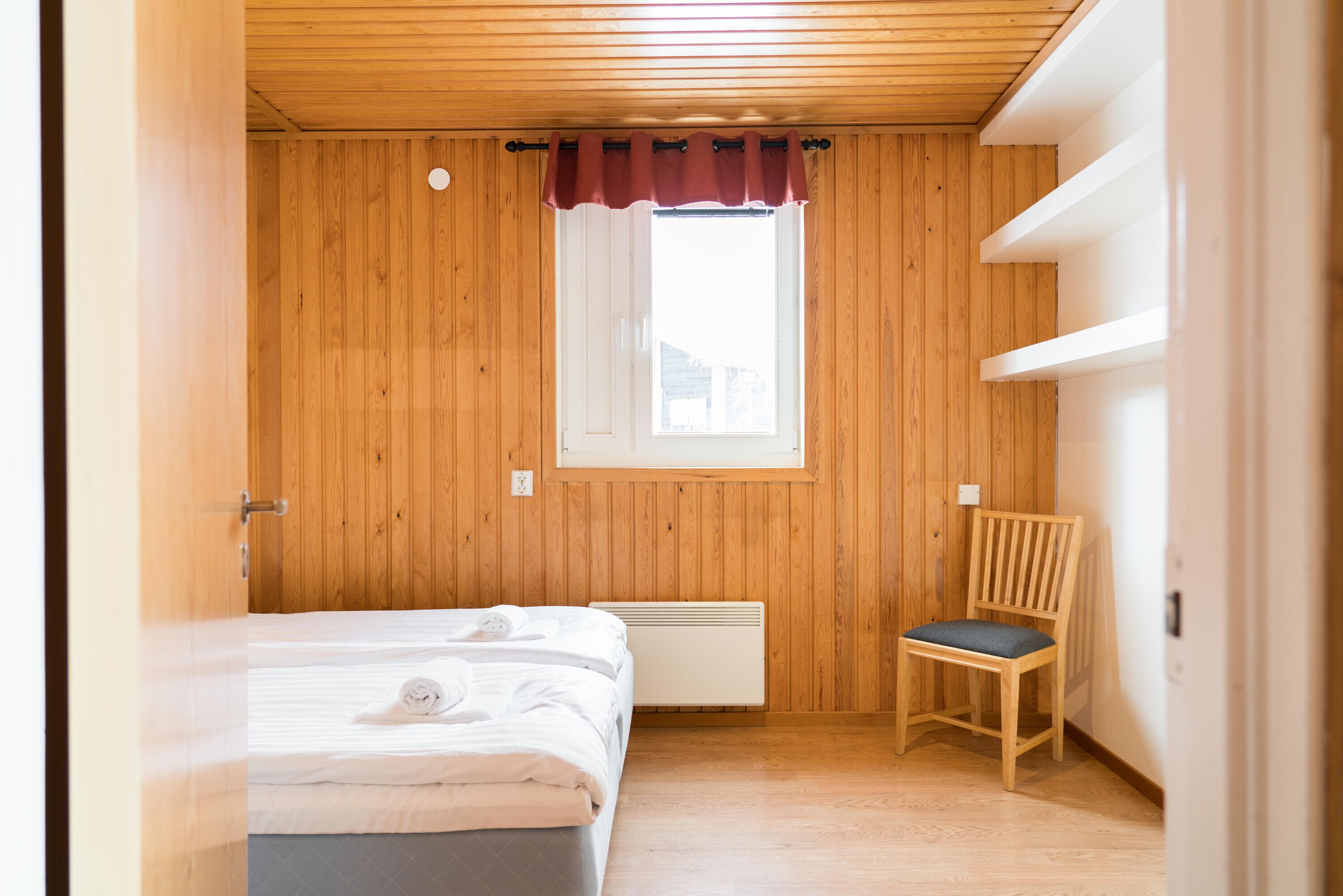 Une chambre avec un mur en bois, une fenêtre ornée d’un rideau rouge, un lit avec une literie blanche, une chaise et une étagère intégrée.
