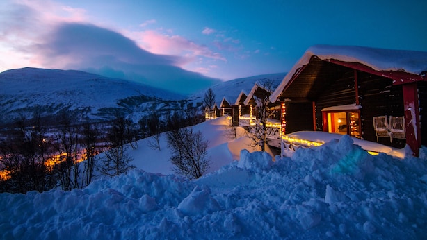 A snow-covered mountain landscape at dusk with a wooden lodge featuring illuminated windows and a snow-covered roof. - Hotell Fjället (Björkliden)