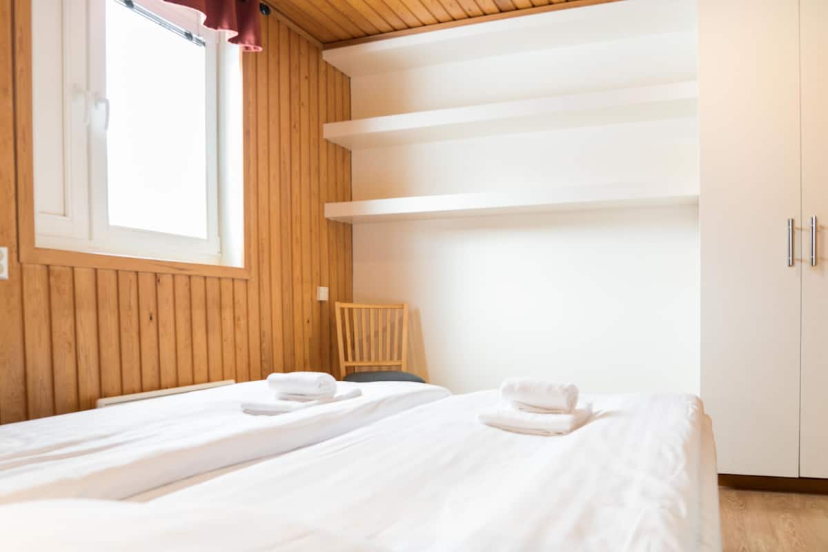 A room with two beds, wooden walls, a window with curtains, and a chair.