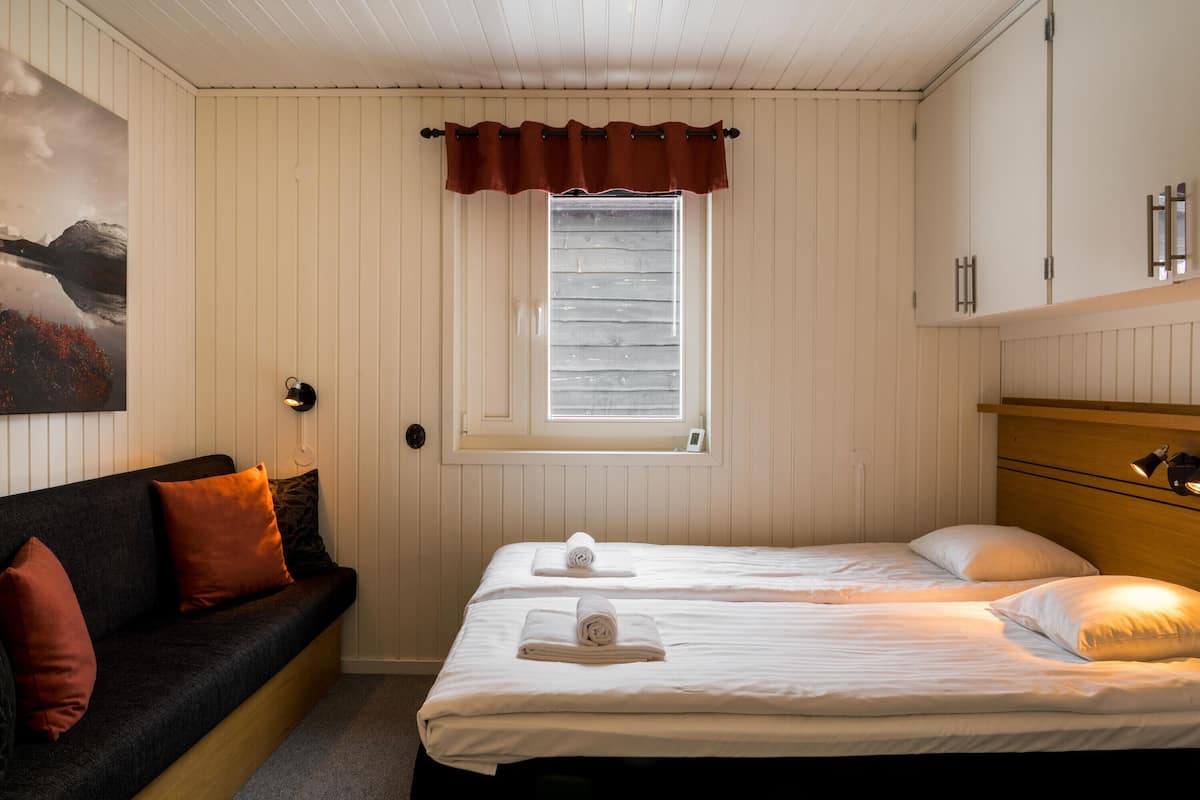A room with two beds, a wooden headboard, a window with blinds, a wall-mounted lamp, and a painting.