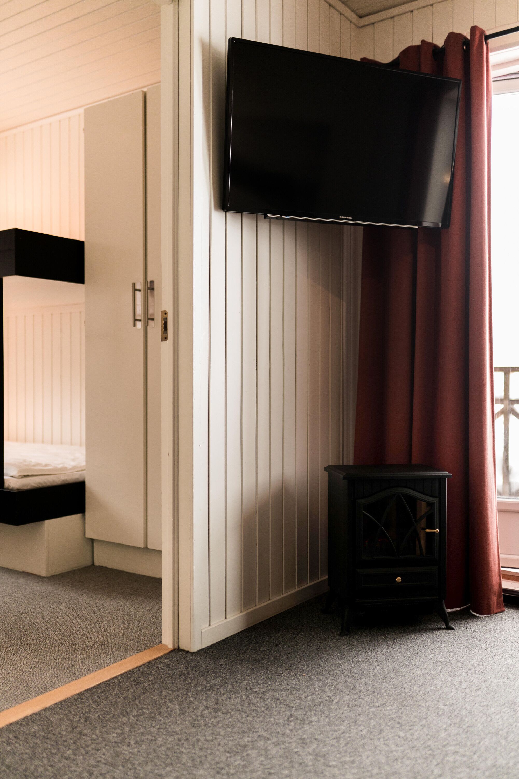 A room with a bed, a TV mounted on the wall, a small black cabinet, and a window with curtains.