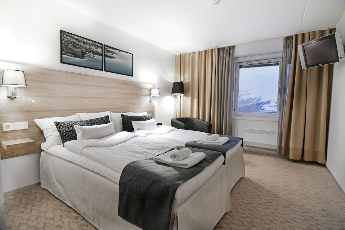 A hotel room with a bed, a TV, a window with curtains, and artwork on the wall.