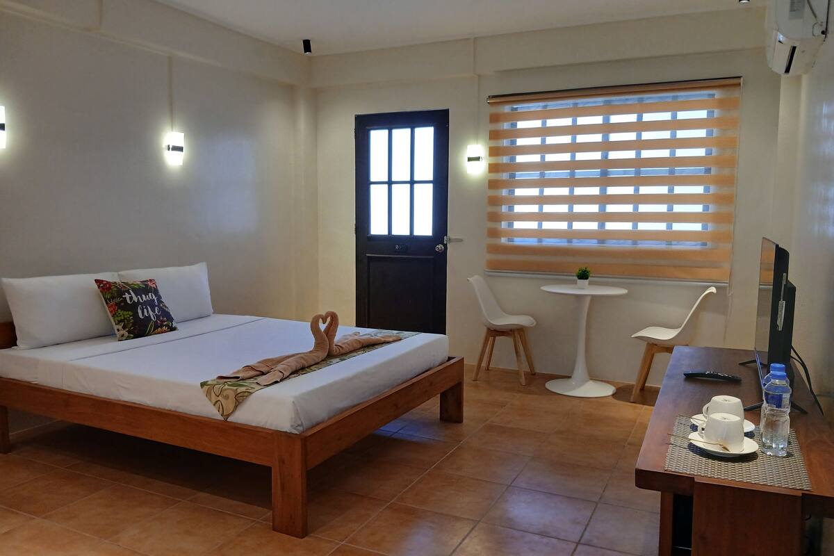 Une chambre d’hôtel comprenant un lit, un bureau avec une chaise, une petite table avec une plante et une fenêtre avec des stores.