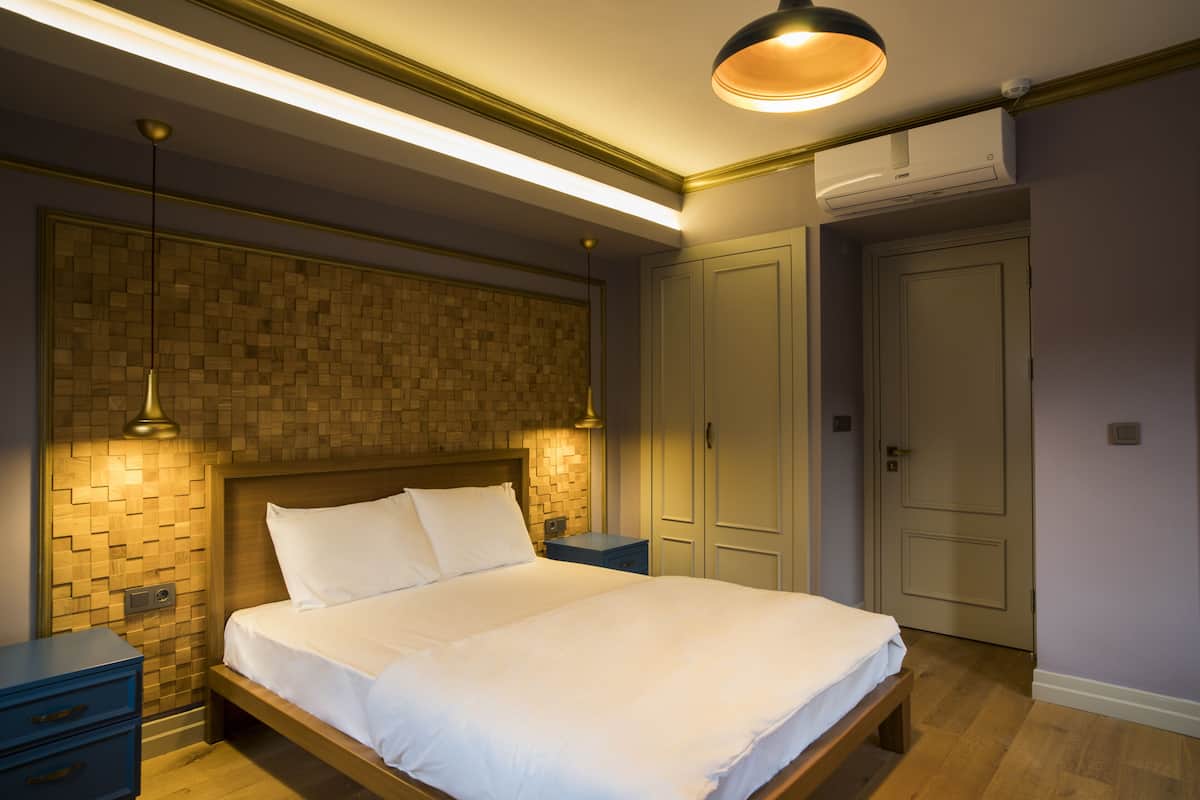 A bedroom with a wooden headboard, a bed with white linens, a nightstand, a blue drawer unit, and a wall-mounted air conditioning unit.