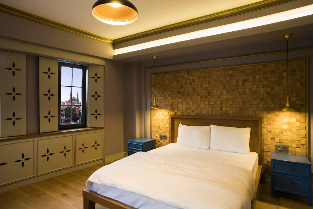 A bedroom with a large bed, a window with a city view, a wooden headboard, and bedside tables.