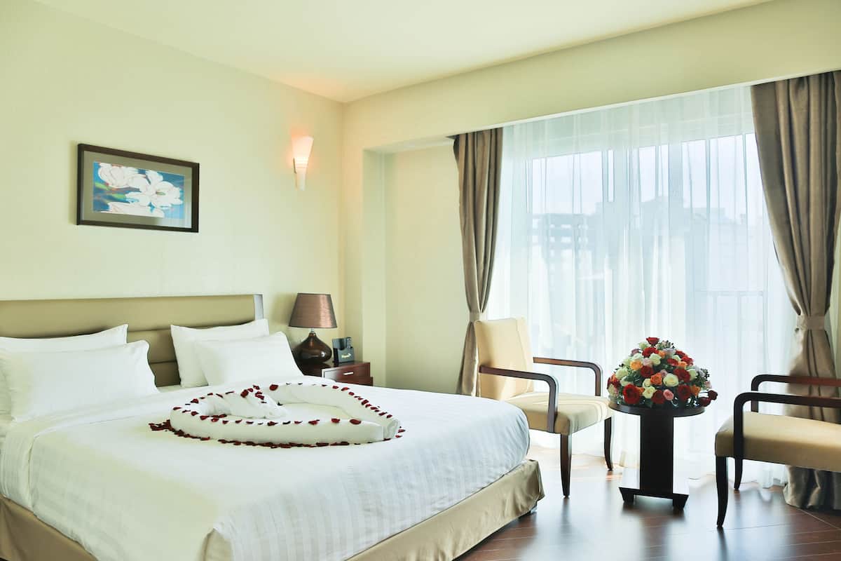 A hotel room with a large bed, a nightstand with a lamp, a chair, a table with a flower arrangement, and a painting on the wall.