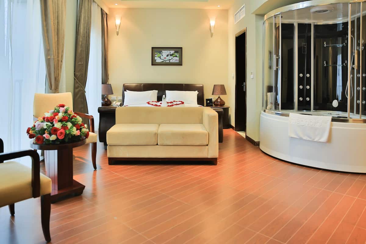 A hotel room with a bed, a sofa, a massage chair, a table with a vase of flowers, and a large window.