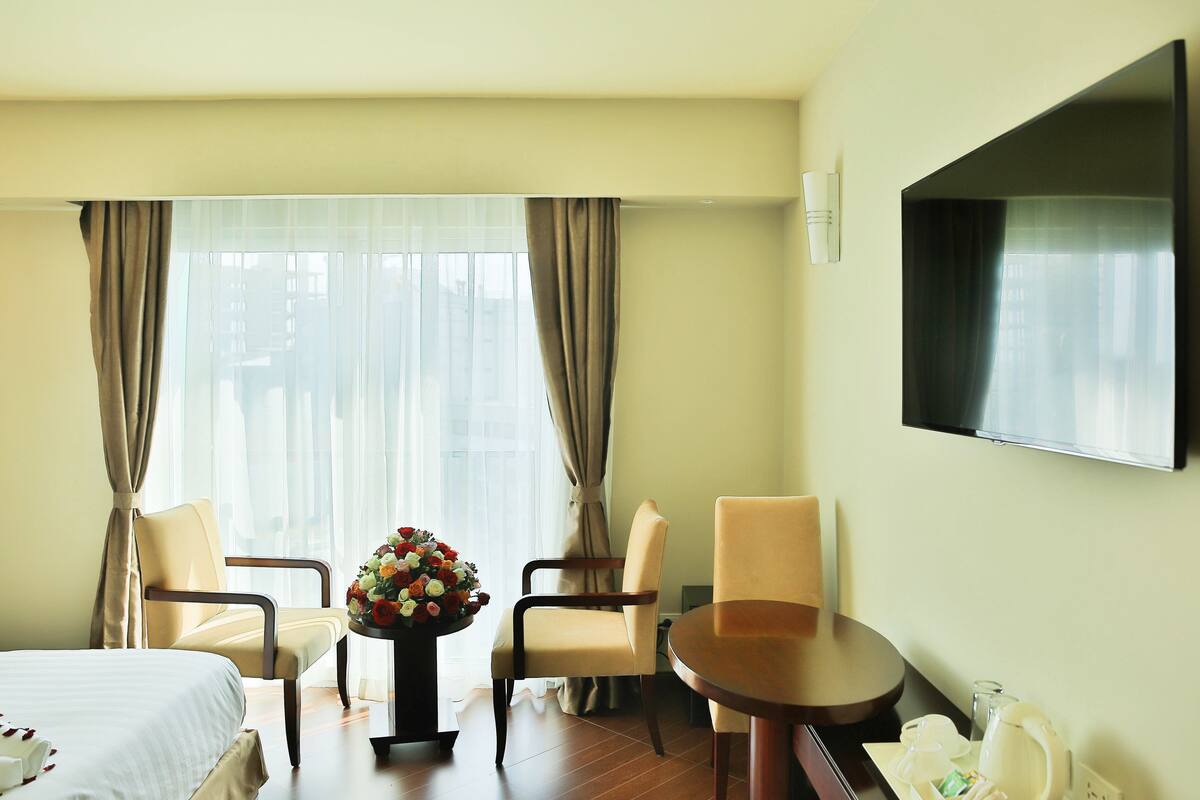 A hotel room with a bed, two chairs, a small table with a vase of flowers, a television, and a window with curtains.