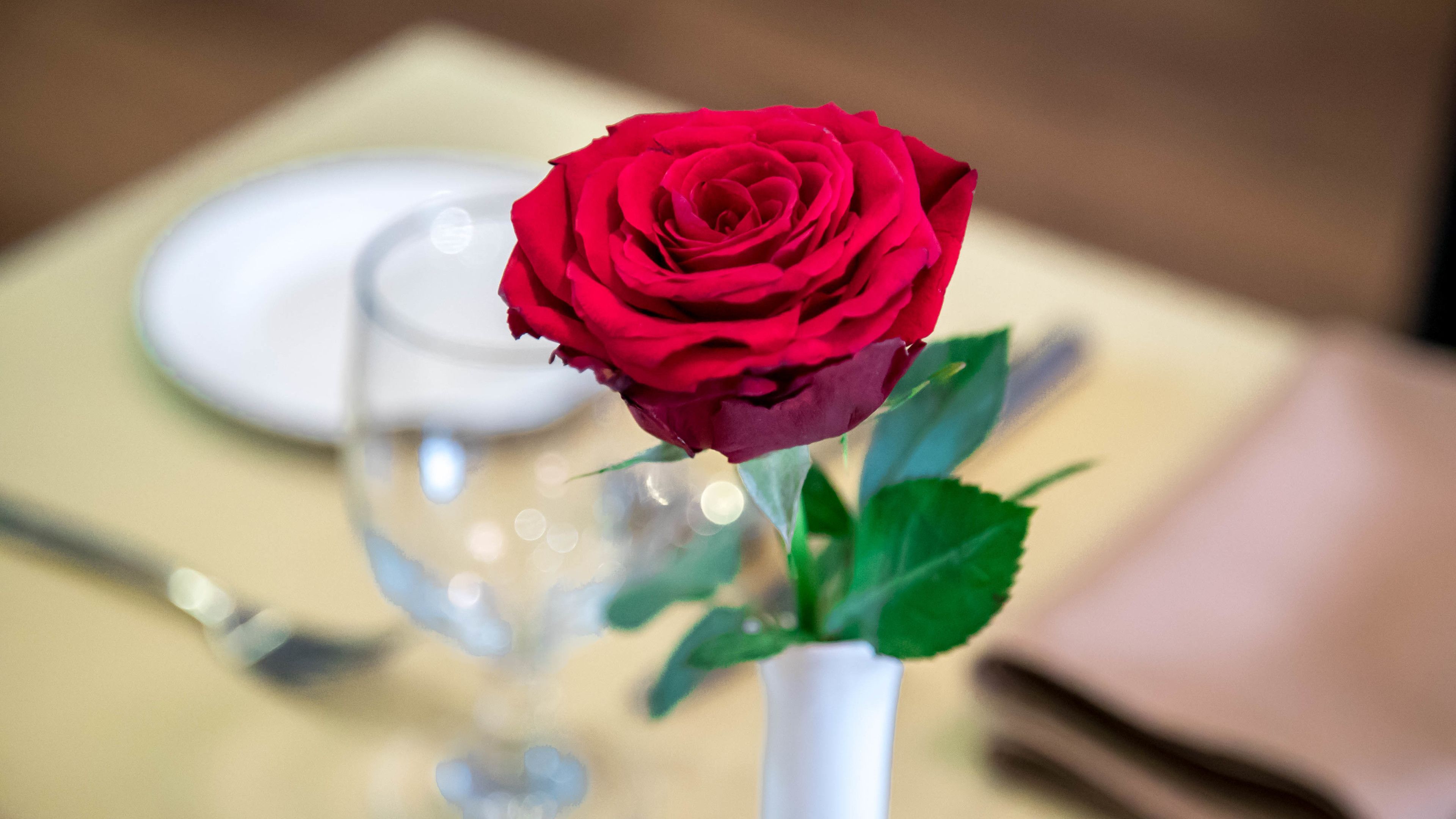 A red rose in a white vase on a table with a plate and a napkin.