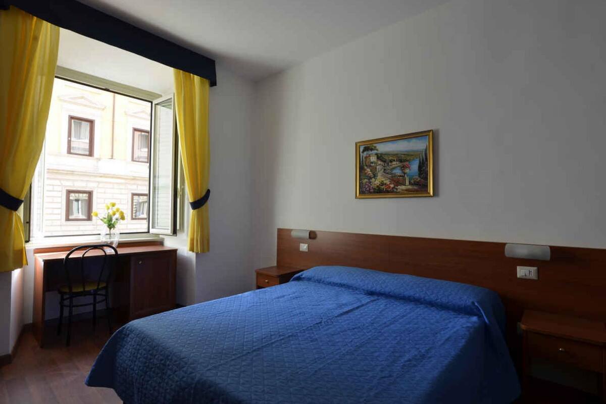 A hotel room with a bed, a desk with a chair, a window with curtains, and a painting on the wall.