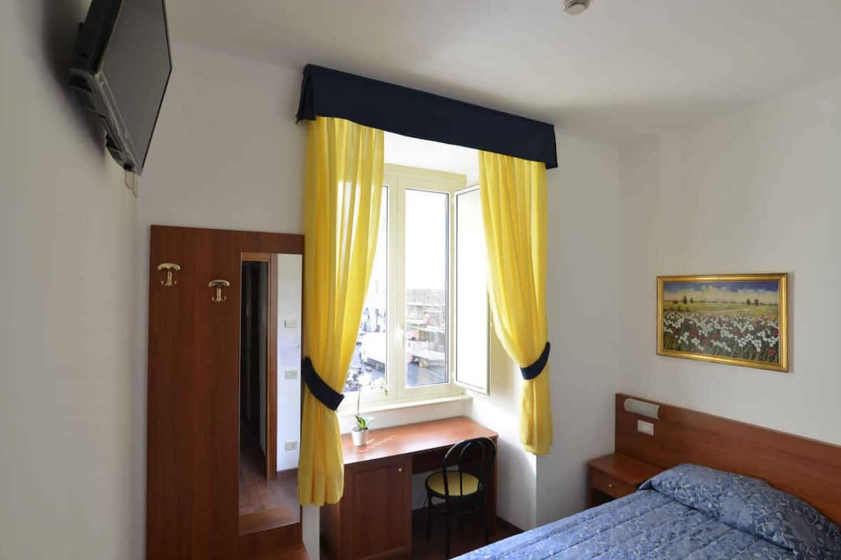 A bedroom with a bed, a desk, and a chair. There is a window with yellow curtains and a painting on the wall.