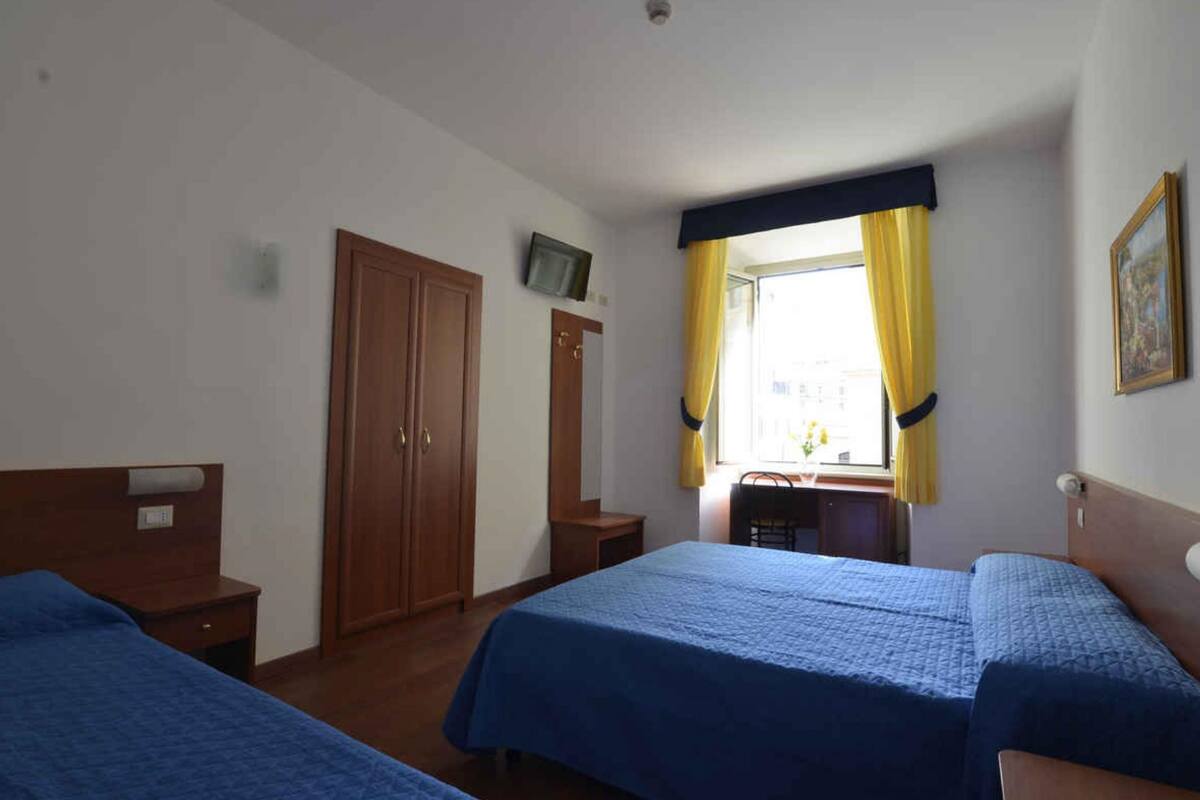 A hotel room with two beds, a wooden wardrobe, a television on the wall, and a window with yellow curtains.