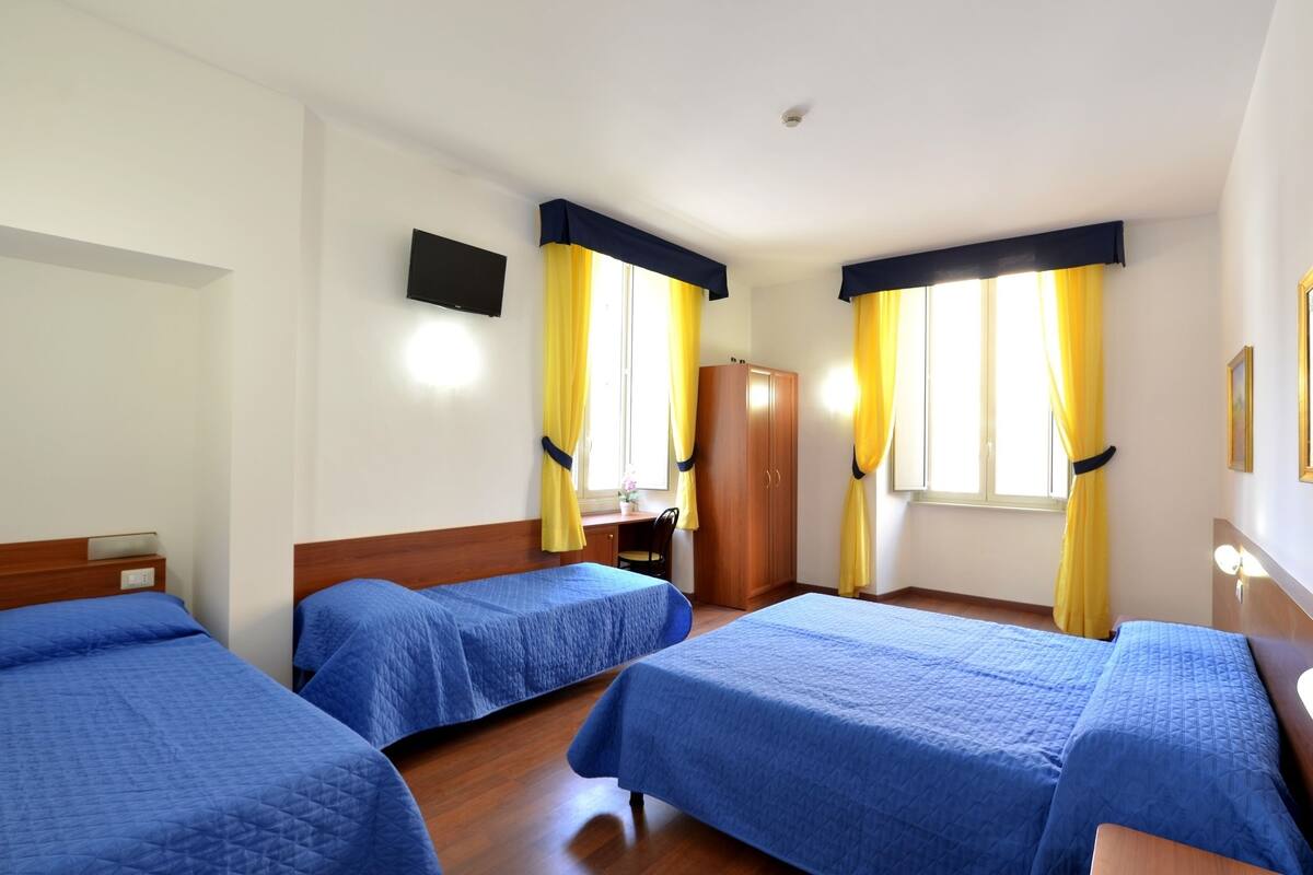 A hotel room with two single beds, a wooden wardrobe, a desk with a chair, and a TV mounted on the wall.