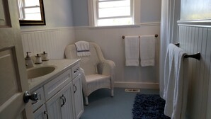 Un baño con tocador blanco, silla de mimbre, toallas blancas y una alfombra azul.
