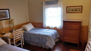 A bedroom with a single bed, a wooden dresser, a chair, a table with a lamp, and a window with lace curtains.