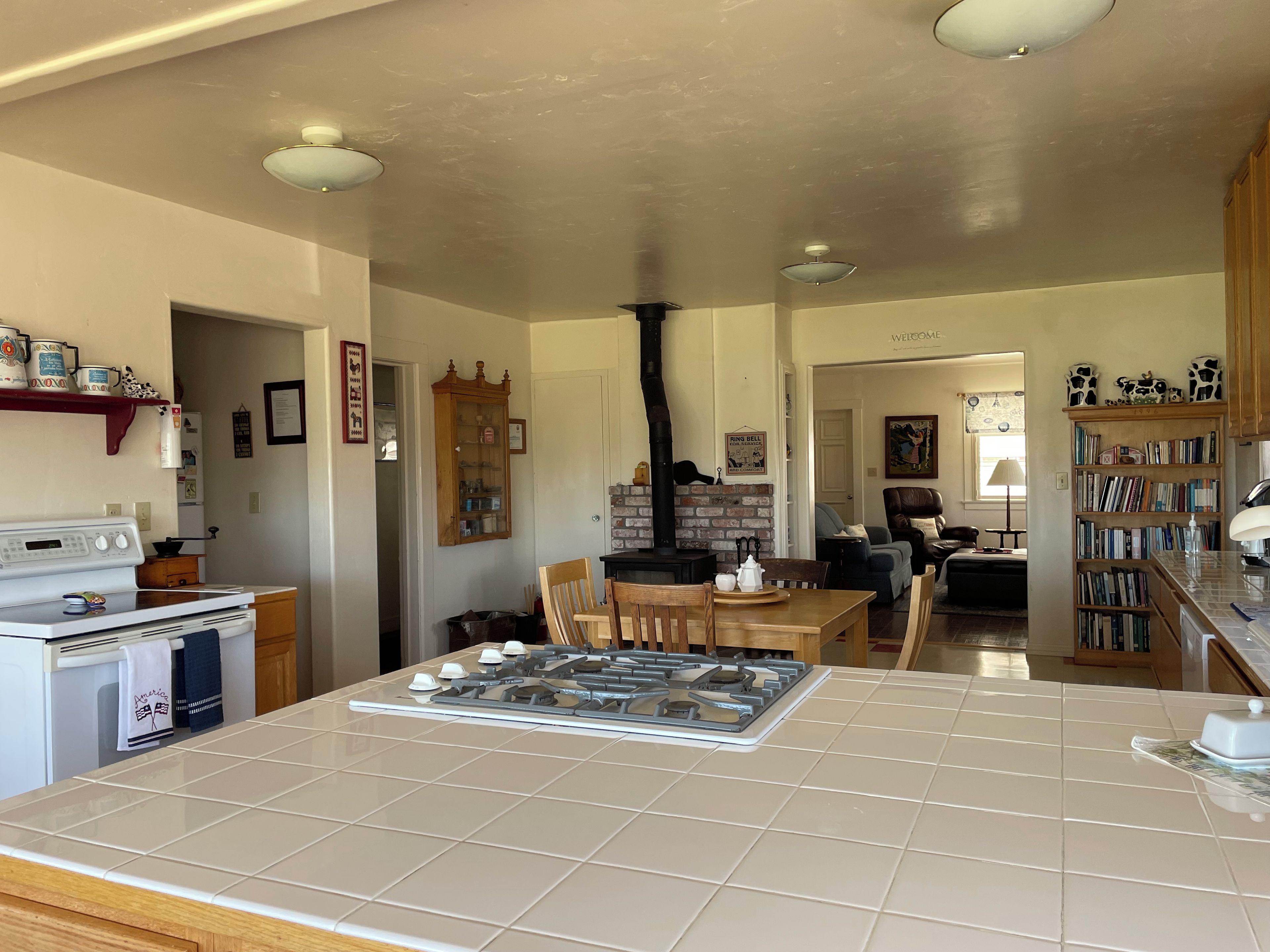 A kitchen with a gas stove, oven, and a brick wall. There is a dining area with chairs and a table, and a bookshelf filled with books.
