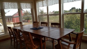 Un comedor con mesa y sillas de madera, ventanas amplias con cortinas de encaje y vistas a un barrio residencial.