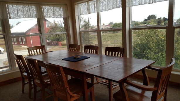 Un comedor con mesa y sillas de madera, ventanas amplias con cortinas de encaje y vistas a un barrio residencial.