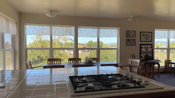 A kitchen with a gas stove, dining table, chairs, and large windows offering a view of greenery and buildings.