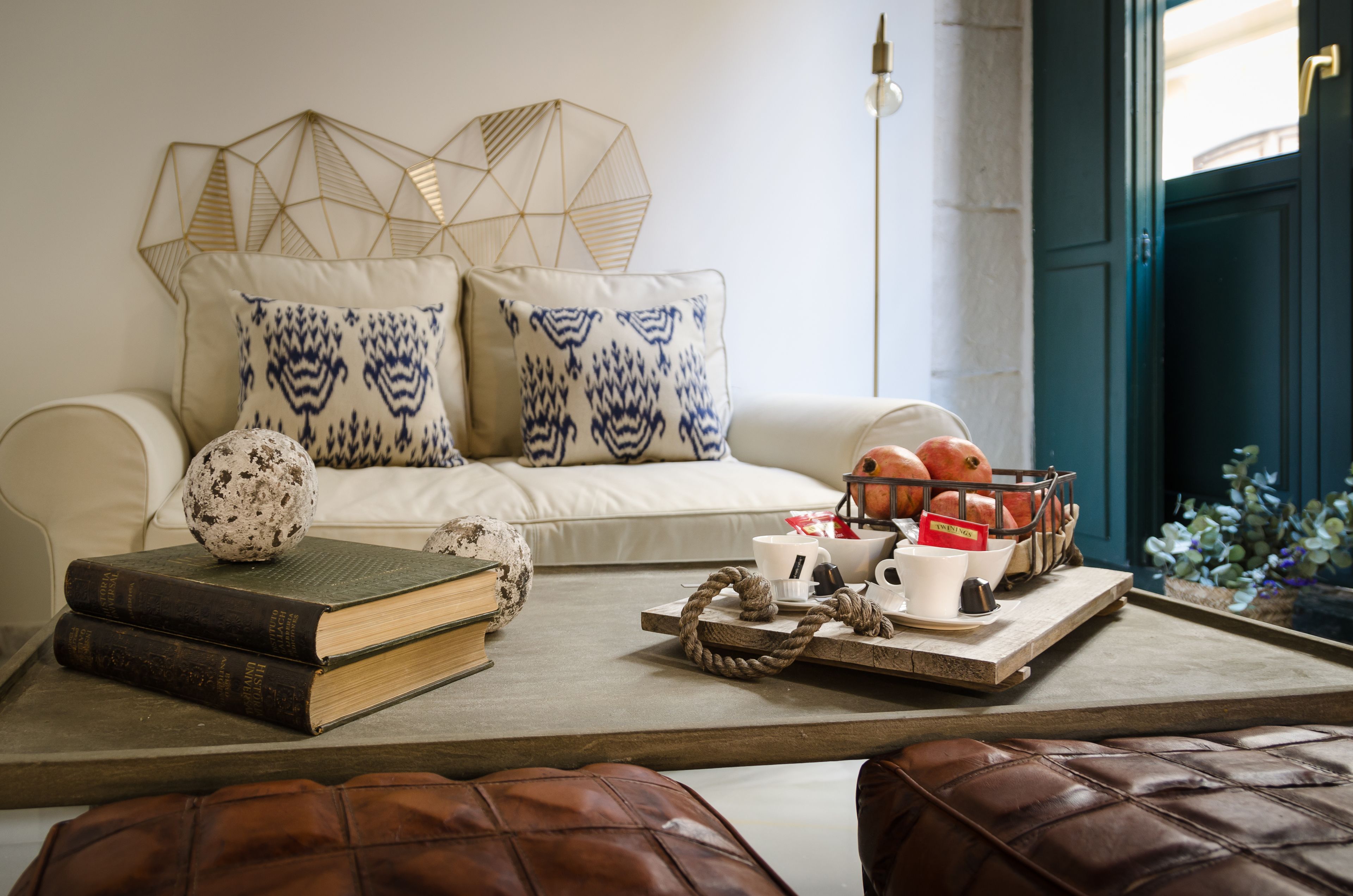 Ein gemütliches Wohnzimmer mit einer Couch, Kissen, einem Couchtisch mit Büchern und einer Ablage sowie einer Tür im Hintergrund.