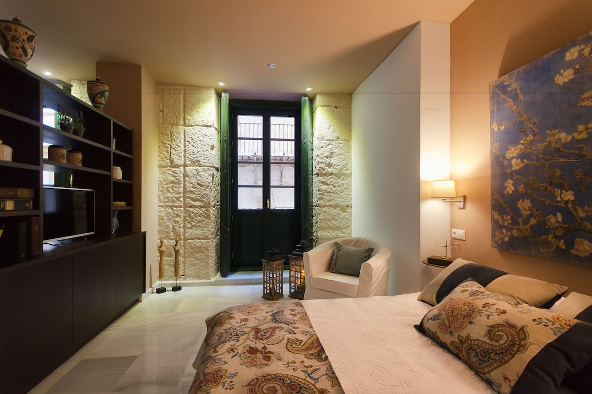A bedroom with a bed, a chair, a TV on a shelf, and a stone wall.