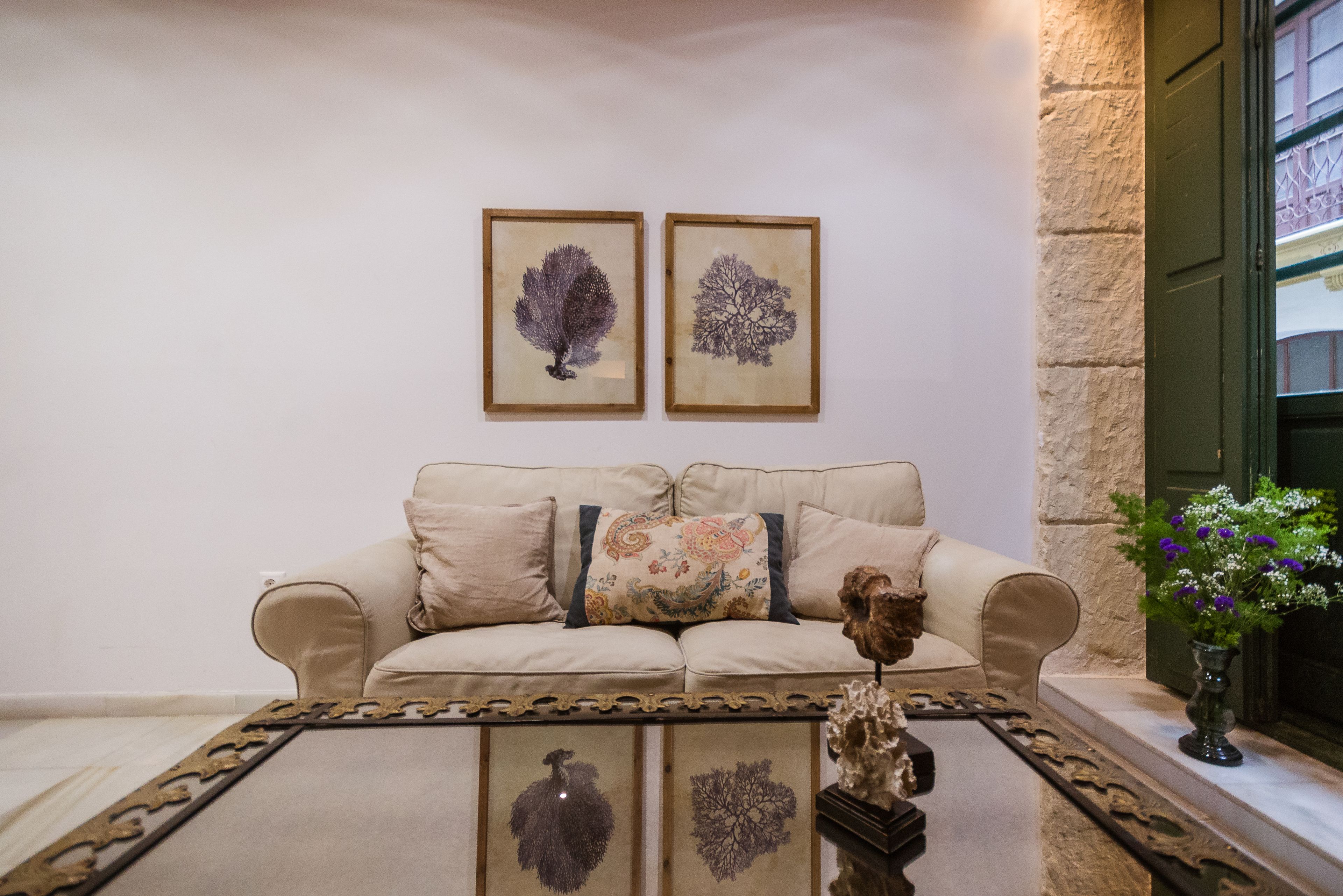 Un salon avec un canapé beige, deux reproductions botaniques encadrées accrochées au mur, une table basse agrémentée d’un objet décoratif et un vase avec des fleurs posé sur un rebord de fenêtre.