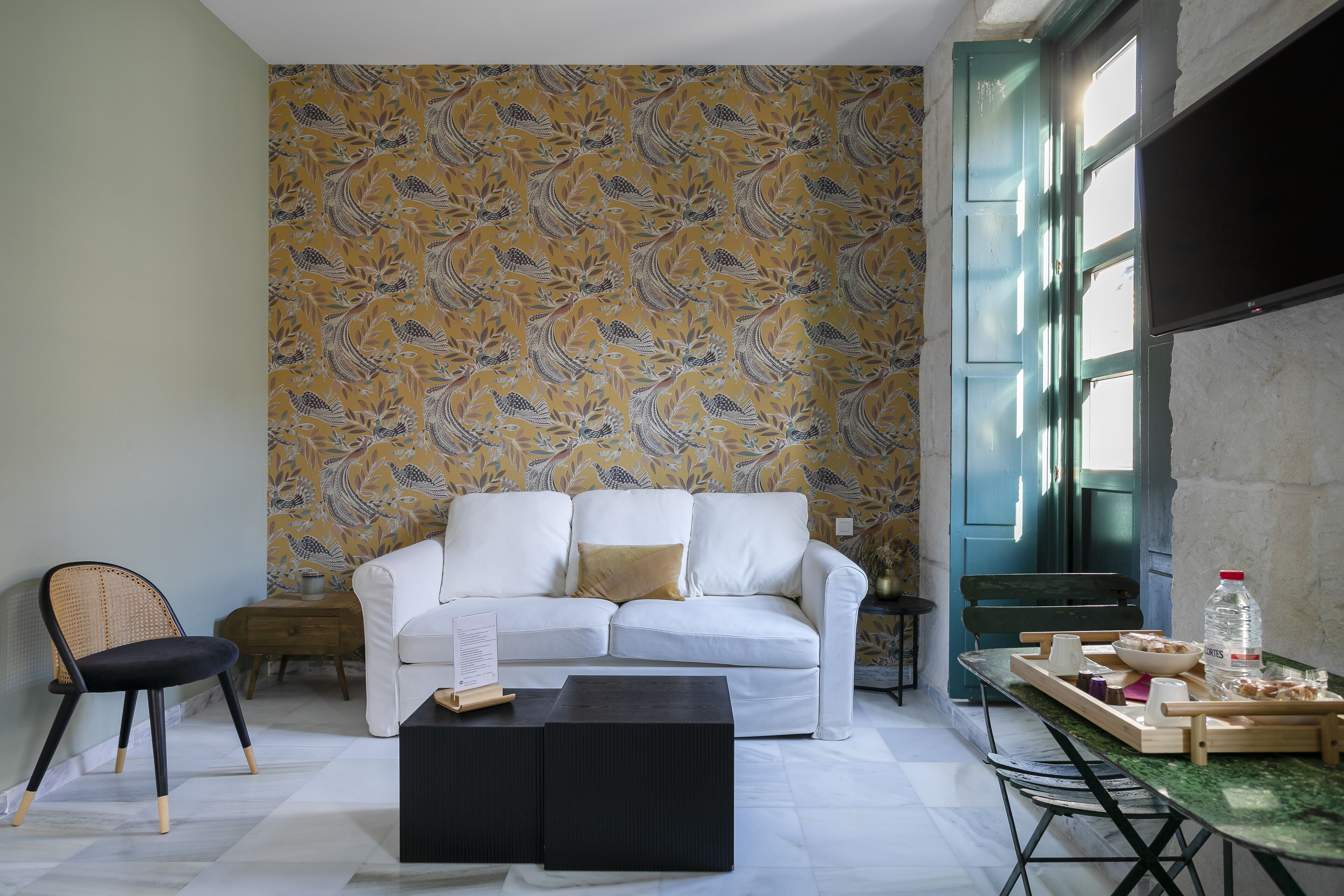 A living room with a white sofa, a black coffee table, a dining table with a tray of food, and a patterned wallpaper.
