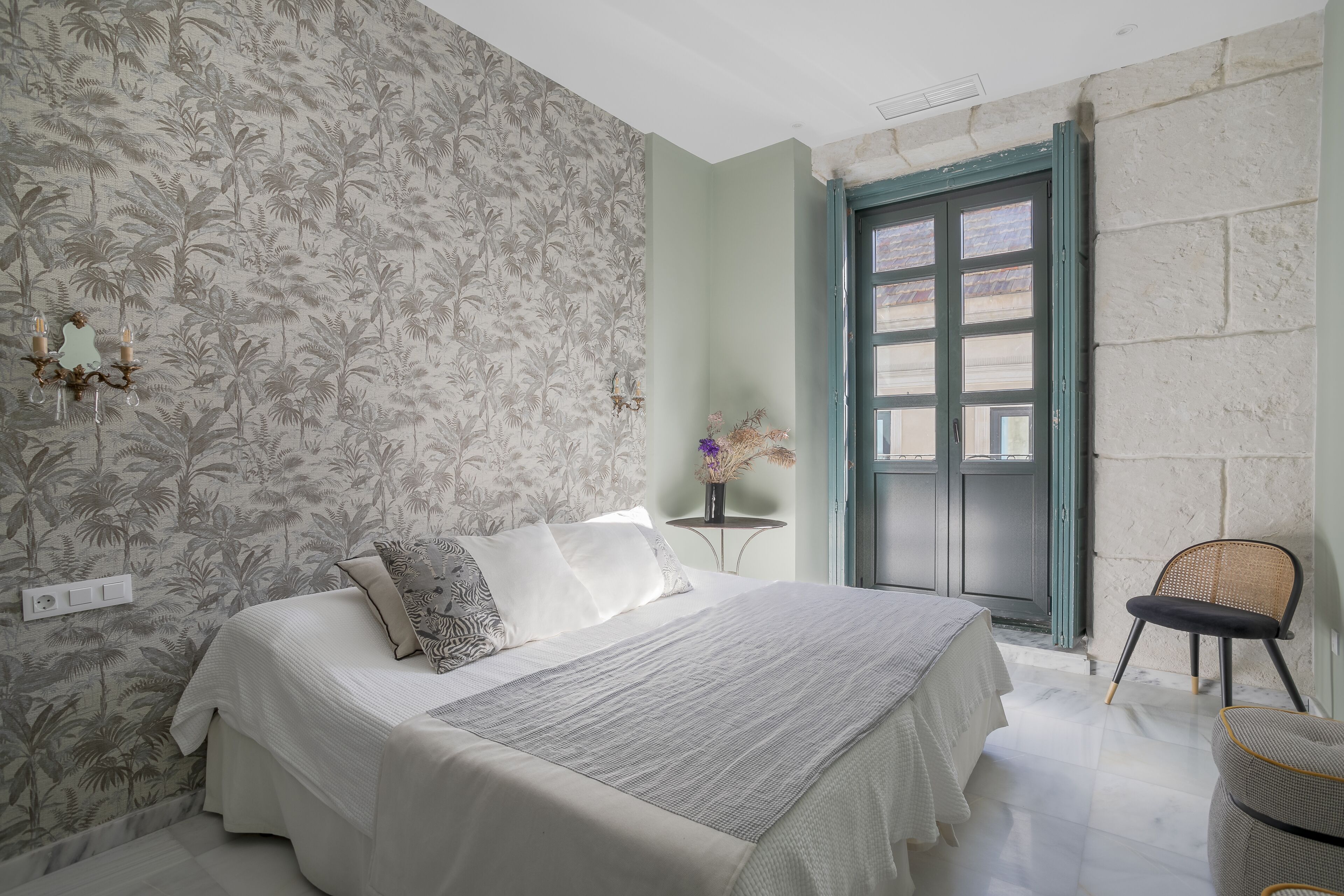 Un dormitorio con cama, mesita de noche, silla, ventana con vistas y pared con estampado floral.