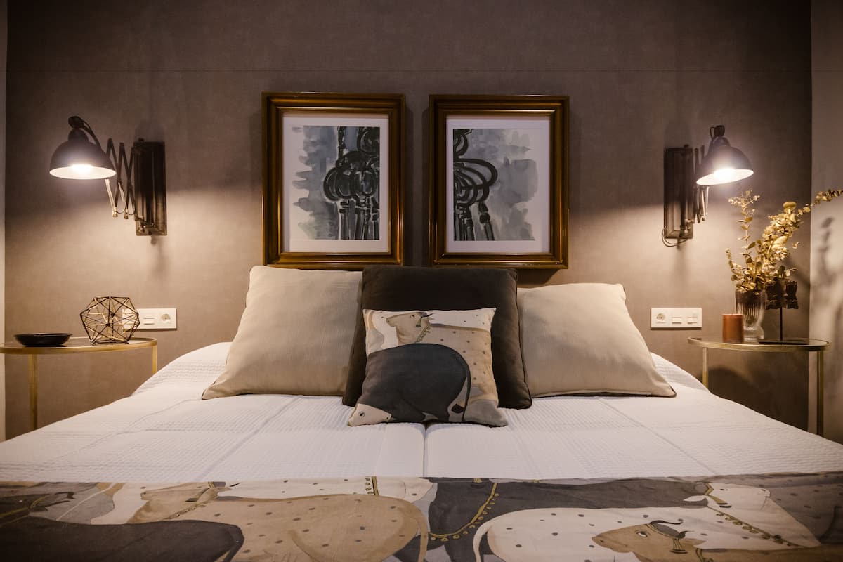 A neatly made bed with two pillows, a headboard, and a bedside table with a lamp and a vase of flowers.