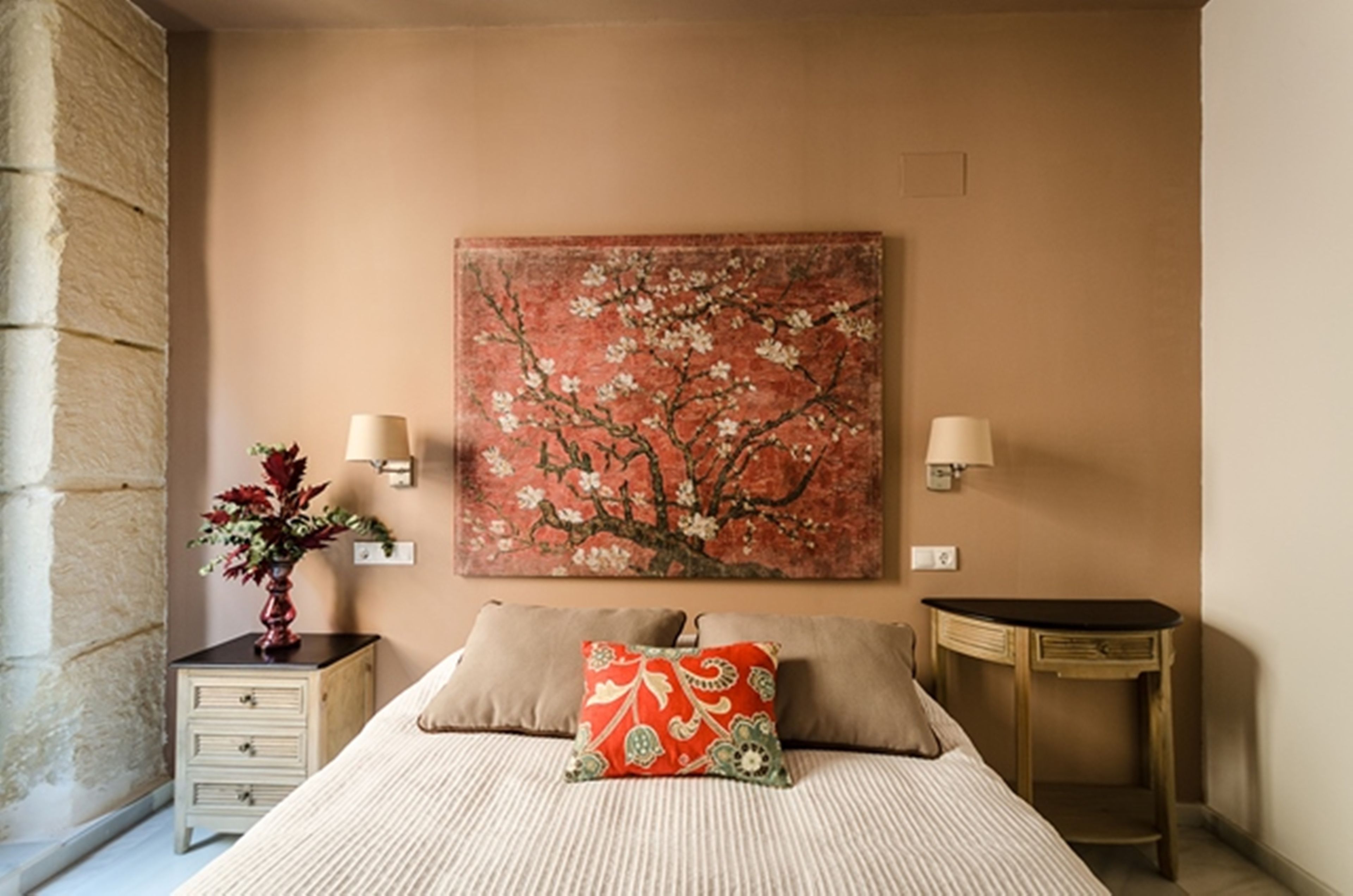 A bedroom with a bed, two bedside tables, a nightstand, a vase with flowers, and a wall-mounted painting.