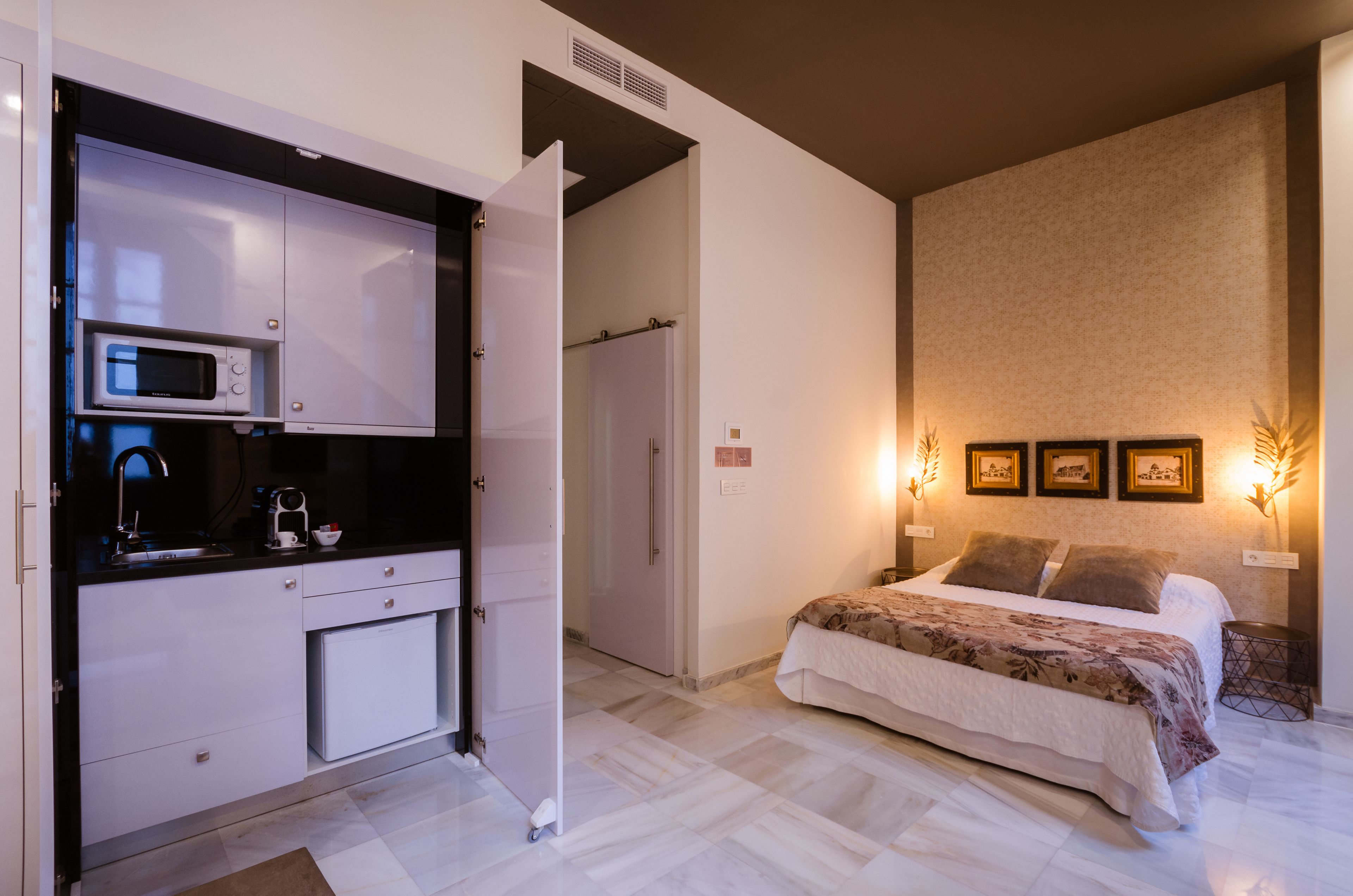 Un dormitorio con cama, mesita de noche, televisor montado en la pared y una zona de cocina con microondas.