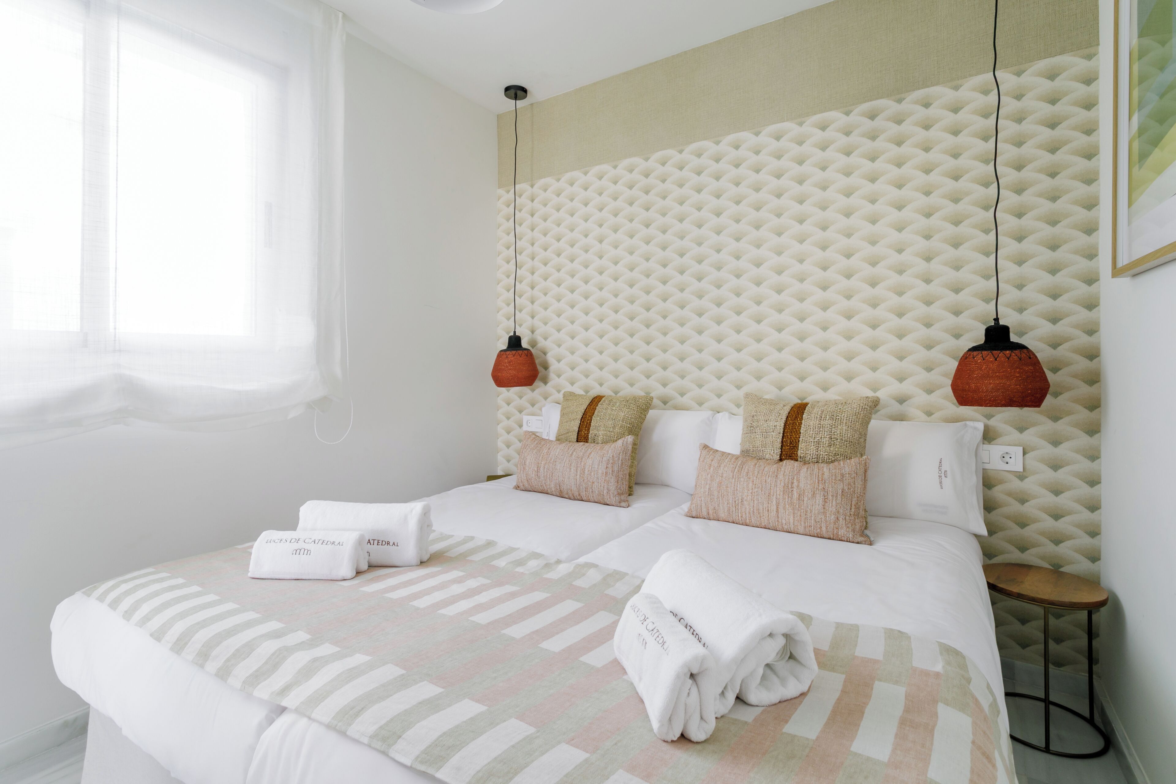 Habitación de hotel con dos camas, un ventanal grande, un muro con textura y dos lámparas colgantes.