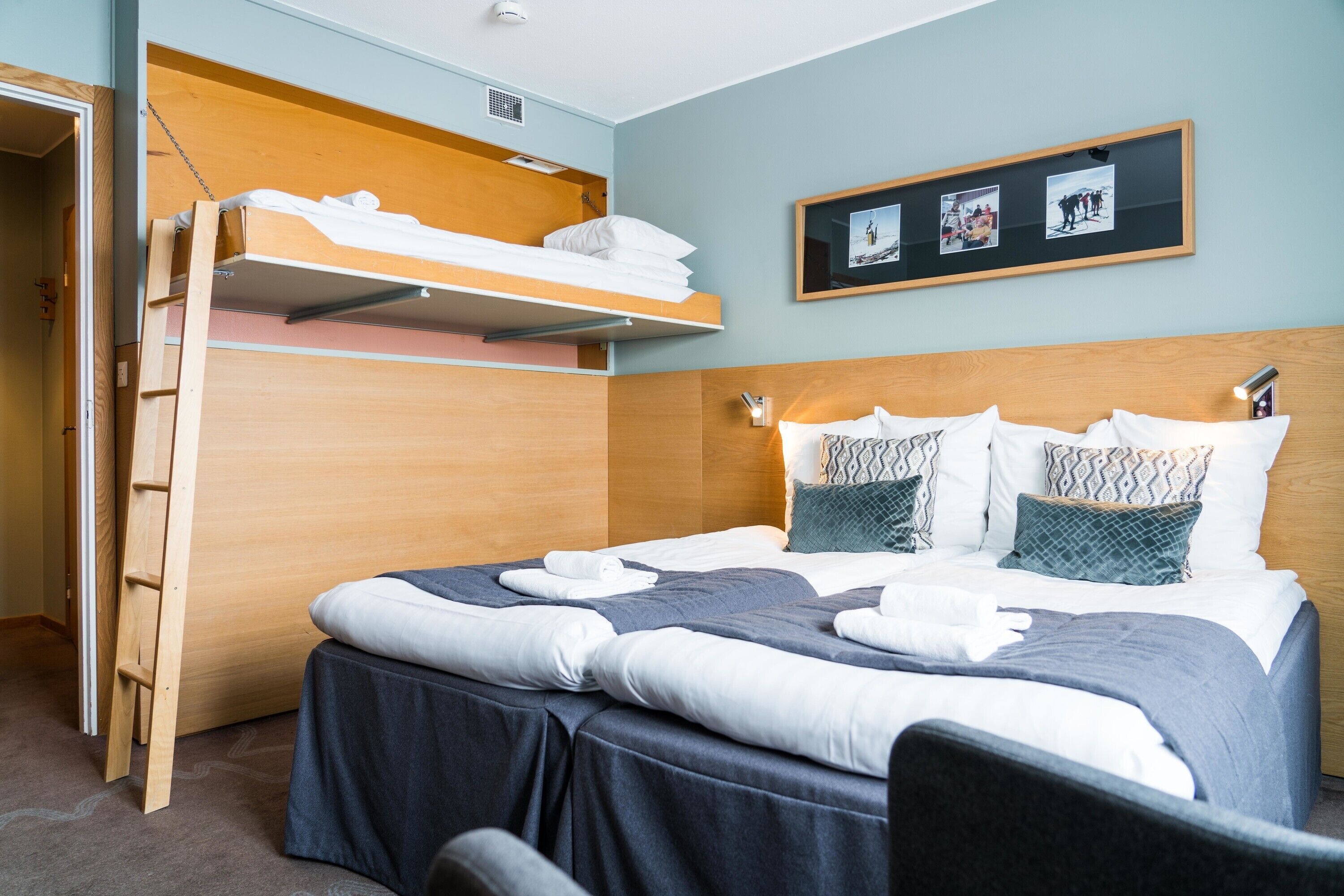 A hotel room with a bunk bed, a desk, and a headboard with a shelf.